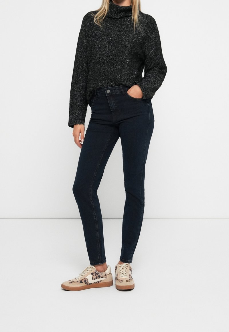 Femme portant un pull à col roulé gris foncé, un jean skinny foncé et des baskets beige à motif léopard, debout avec une main dans la poche.