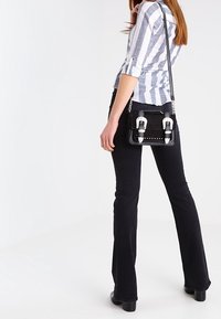 Donna con pantaloni neri a zampa d'elefante e camicia a righe che porta una borsa nera con fibbie argentate sulla spalla, in piedi davanti a uno sfondo bianco.