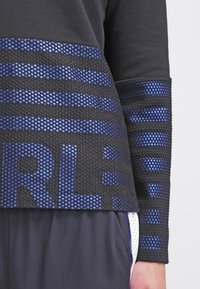 Top deportivo negro con textura de malla, acentos a rayas azules y un diseño gráfico audaz "RL". Mangas largas con patrones contrastantes.