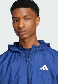 Mørkeblå letvægtsjakke med lynlås og hætte. Har hvidt Adidas-logo på brystet. Glat overflade, moderne design.