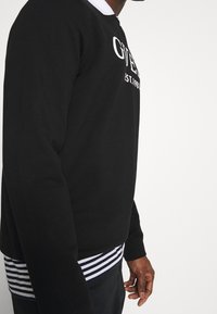 Sudadera negra con logo blanco de "Guess", puños acanalados y detalle de dobladillo a rayas. Presenta un cuello redondo y un ajuste relajado.