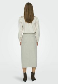 Lichtgrijze jurk van gemiddelde lengte met achterzakken, gecombineerd met een crèmekleurige blouse met bloemborduursels op de schouders. Donkerbruine enkellaarsjes.