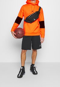 Felpa arancione con accenti neri, marsupio nero, pantaloni corti da basket neri e sneakers nere con strisce grigie. Modello che tiene un pallone da basket.