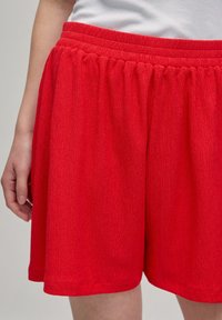 Rote, strukturierte Shorts mit elastischem Bund. Der Stoff hat ein dezentes Muster, endet über dem Knie und fällt locker.