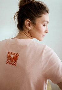 Hellrosa Baumwoll-T-Shirt mit einem orangefarbenen Schachbrett-Grafik und Fruchtillustrationen auf der Rückseite. Kurze Ärmel und lockere Passform.