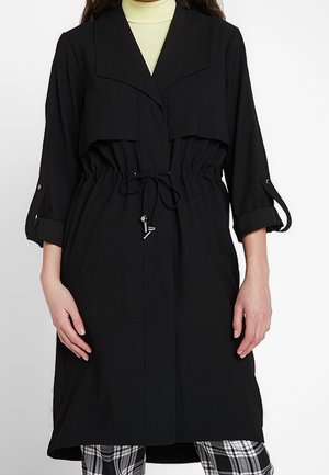 Zwarte trenchcoat met lange mouwen en opgerolde manchetknopen, verstelbare taille met trekkoord, gedragen over een lichte coltrui en geruite broek.