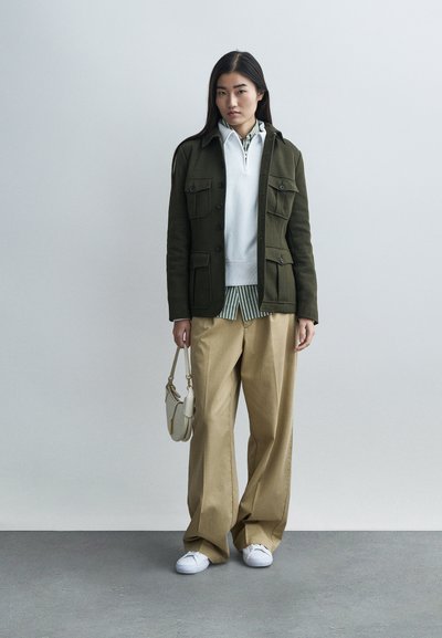 Olivno zelena jakna z žepi, bela jopa z zadrgo, črtasta majica, široke bež hlače in beli supergi; drži majhno kremo torbico.