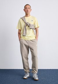 Šviesiai geltona marškinė su mėlynu logotipu, béžiniai sportiniai kelnių, ir pilki sportiniai batai. Megztinis per pečius, stovintis ant mėlynos kilimo.