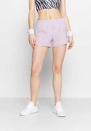 Kvinde iført lyse lilla træningsshorts, sort-hvidt zebraprint crop top, hvide håndledsbånd og hvide sneakers, som står mod en hvid baggrund.