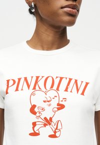 Κοντινό πλάνο ατόμου που φορά λευκό πουκάμισο με πορτοκαλί κείμενο "PINKOTINI" και μια καρδιά που χορεύει κρατώντας ένα ποτήρι μαρτίνι και μουσικές νότες.