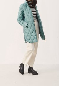 Chaqueta larga acolchada en verde menta con cremallera frontal, bolsillos laterales y cuello alto, combinada con botas negras gruesas y pantalones crema.