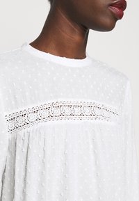 Blusa bianca con texture a pois e inserto in pizzo sul petto. Tessuto morbido con scollatura rotonda e design arricciato.