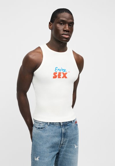 Λευκό βαμβακερό αμάνικο τοπ με το κείμενο "Enjoy SEX" σε μπλε και κόκκινο, συνδυασμένο με ανοιχτό μπλε τζιν σορτς με φθαρμένη εμφάνιση και κεντημένες λεπτομέρειες.