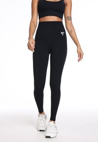 Zwarte high-waisted legging met een gladde textuur en een klein wit logo op de linkse heup. Gekenmerkt met een zwarte crop top en witte sneakers.