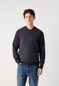 Ναυτική bomber ζακέτα με μπροστινό μέρος από λείο ύφασμα, πλεκτά μανίκια, ριμπ μανσέτες και λογότυπο στο στήθος. Φοριέται με μπλέ τζην παντελόνι.