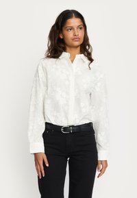 Weißes, blumengemustertes Langarmhemd mit Button-Down-Kragen, kombiniert mit schwarzen Jeans und einem schwarzen Gürtel mit silberner Schnalle.