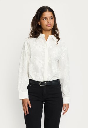 White floral-patterned long-sleeve shirt with a button-down collar, paired with black jeans and a black belt with a silver buckle.