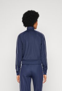 Giacca di tuta blu navy con colletto alto, strisce bianche ai lati, polsini a coste e silhouette aderente, realizzata in un tessuto liscio e elastico.