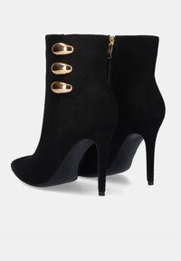 Botines negros de ante con punta afilada, altos tacones de aguja, tres acentos decorativos en tono dorado y cierre de cremallera lateral.