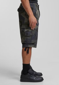 Kamouflage cargo shorts i mörkgrön och svart, med flera fickor, knälång design och sidofästen. Bärs med svarta sneakers och strumpor.
