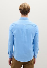 Camicia azzurro chiaro a maniche lunghe con colletto, piega posteriore e polsini con bottoni, realizzata in tessuto liscio, abbinata a pantaloni marroni.