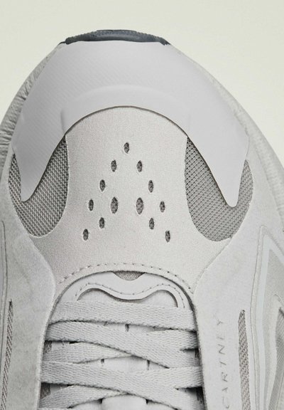 Chaussure de sport gris clair en mesh et cuir suédé. Présente une tige texturée, des détails en lacets et des trous de ventilation près des orteils.