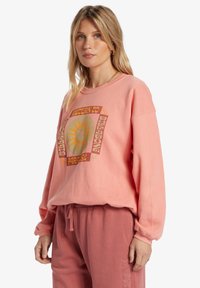 Sweatshirt rose clair à manches longues, avec un design graphique de soleil en orange et jaune à l'avant. Texture douce, coupe décontractée.