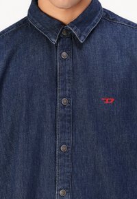 Dark denim shirt with a button-down collar, featuring silver buttons and a small red logo on the left side. Textured fabric with subtle fading.