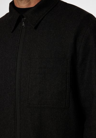 Svart ullblandad jacka med dragkedja, spetsig krage och en bröstficka med en något texturerad yta.