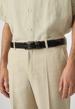 Mann trägt beige strukturierte Hose und ein hellbeiges Hemd mit Knöpfen sowie einen schwarzen Ledergürtel in der Taille.