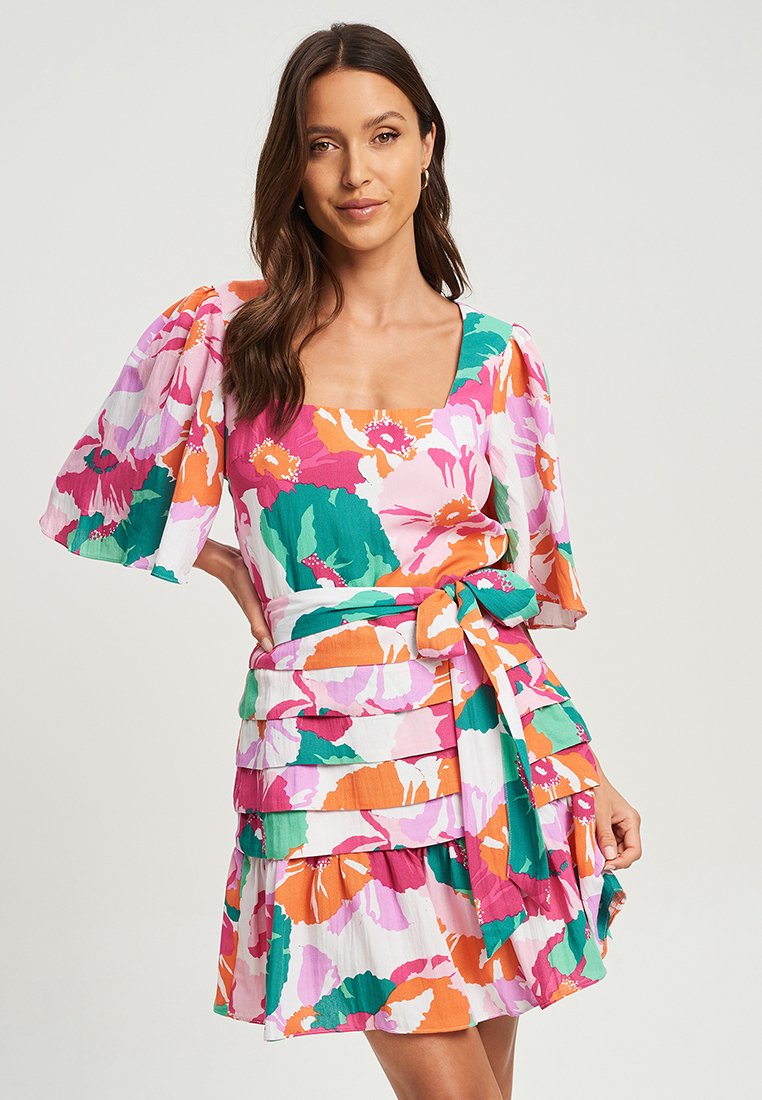 Vrouw met lang bruin haar, gekleed in een kleurrijke bloemenjurk met poffende mouwen, vierkante halslijn en een gelaagde rok die bij de taille is vastgebonden.