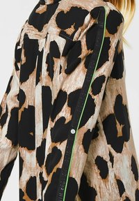 Camicia con stampa leopardata in tonalità marrone e nera, realizzata in tessuto testurizzato, con piping verde a contrasto e un dettaglio a bottoni lungo la cucitura laterale.