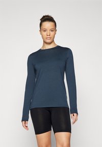 Long-sleeve shirt in dark blue fabric with a round neck, smooth texture, and a small logo on the chest; paired with black shorts.