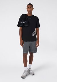 Jonge man in een zwart Champion t-shirt, grijze shorts, witte sokken, grijze sneakers, met een zwart Champion handdoek over zijn schouder.