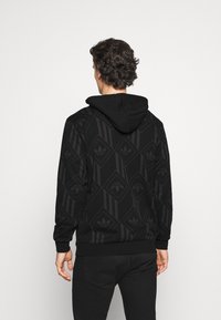 Černá mikina s kapucí s texturovaným diamantovým vzorem a třemi pruhy po stranách. Má kapuci se stahovací šňůrkou a žebrované manžety.