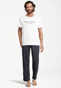 Weißes Baumwoll-T-Shirt mit schwarzem Druck "Marc O’Polo EST. IN STOCKHOLM", kombiniert mit navyblauen Hosen, glatte Textur, lockere Passform und gerade Beine.