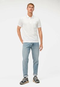 Witte polo met een zak, korte mouwen en een contrasterende kraag. Draagt een lichtblauwe spijkerbroek en zwarte sneakers met beigekleurige accenten.