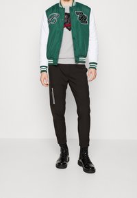 Veste universitaire verte et blanche avec lettres brodées, pull gris avec motif d'ours, pantalon noir fuselé avec poche zippée latérale, et bottes noires.
