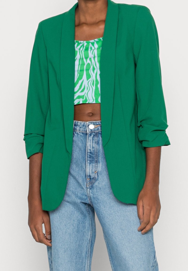 Green blazer with a smooth texture, featuring a tailored fit, notched lapels, and rolled sleeves, paired with a patterned crop top and high-waisted jeans.