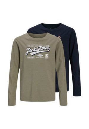 Dreng står foran med front mod kamera iført olivengrøn langærmet skjorte med tekst, løstsiddende blå jeans og beige sko mod en ensfarvet baggrund.
