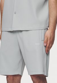 Lichtgrijs geribbeld shirt en shortset, met een knoopdetail op het shirt en een geborduurd logo op de shorts. Zachte textuur.