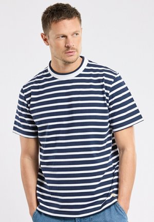 Homme aux cheveux courts portant un t-shirt à rayures horizontales bleu marine et blanc, debout les mains dans les poches, regardant sur le côté.