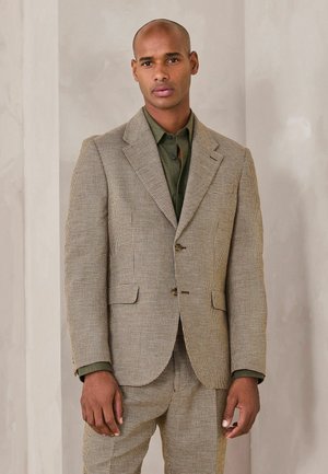 Mann trägt einen braun-schwarz karierten Anzug mit olivgrünem Hemd, steht vor einer schlichten, hellen Wand.