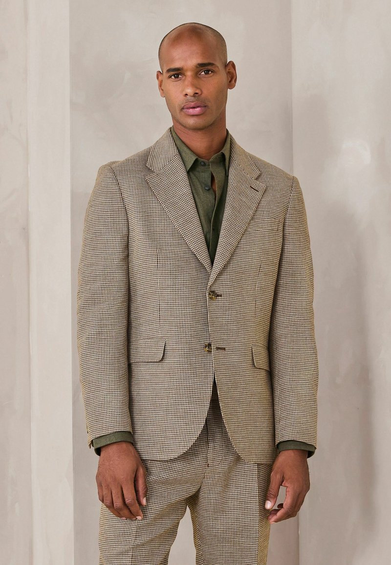 Muž v béžovo-čiernom károvanom obleku s olivovo zelenou košeľou stojaci pred jednoduchou svetlou stenou.