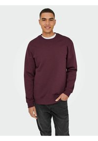 Bordeauxrode sweatshirt met geribbelde manchetten en zoom, ronde halslijn, gemaakt van een zachte stof. Casual gedragen over een wit shirt met donkere jeans.