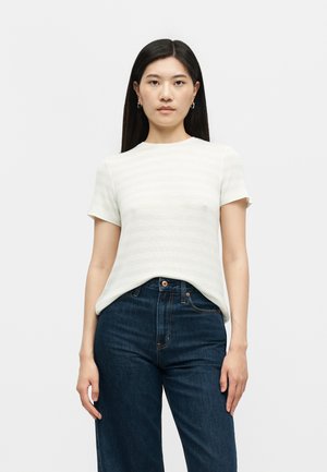 Γυναίκα με μακριά σκούρα μαλλιά, που φοράει λευκό πουκάμισο με κοντά μανίκια και σκούρα μπλε τζιν με ψηλή μέση, στέκεται μπροστά σε απλό ανοιχτό φόντο.