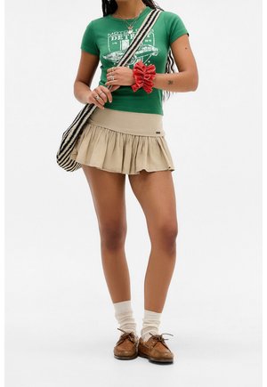 Mujer con camiseta gráfica verde, minifalda plisada beige, calcetines blancos, zapatos marrones, coletero rojo y bolso de hombro a rayas negras y blancas.