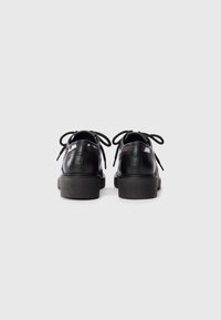 Chaussures en cuir noir avec une finition brillante, semelles plates, bout arrondi, et lacets. Marquage discret sur le talon et l'arrière.