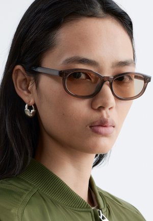 Mujer con gafas de sol rectangulares tintadas en marrón, pendientes de aro plateados y una chaqueta verde oliva con cuello acanalado.