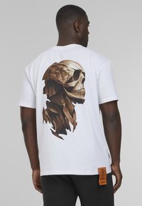 White t-shirt featuring a large, detailed graphic of a wooden skull on the back, with an orange tag on the side seam.